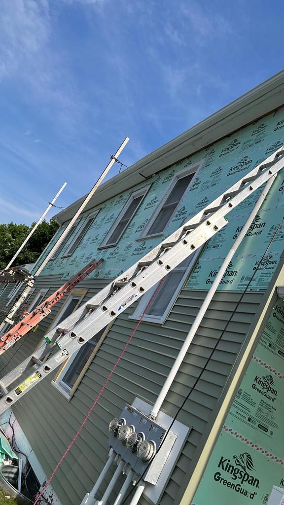 Upward view of a roof eave and gutter line above house-wrap siding, ladder in the foreground
