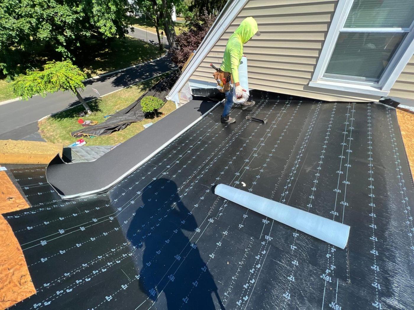 Crew member installing synthetic roofing underlayment on an angled roof beside a dormer window
