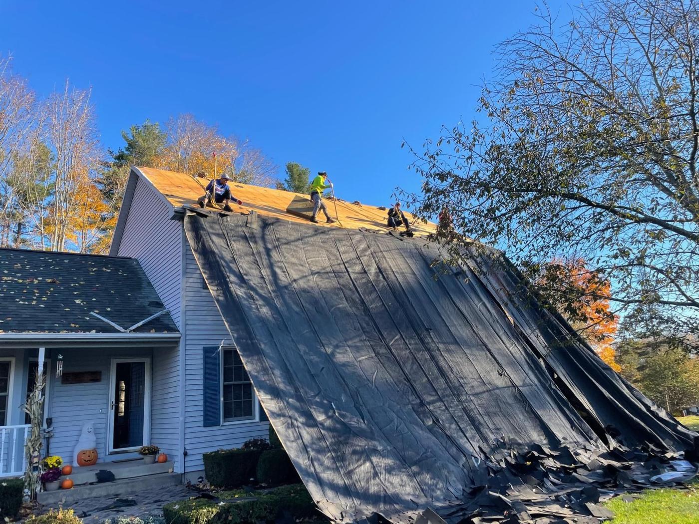Steep A-frame roof with black synthetic underlayment draped down over the tear-off, crew working the ridge, autumn trees behind