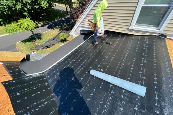 Start Roofing crew member installing synthetic underlayment on a sloped roof beside a dormer window