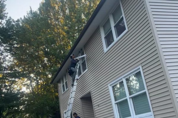 Crew on a ladder working near the roofline of a light-sided home surrounded by trees